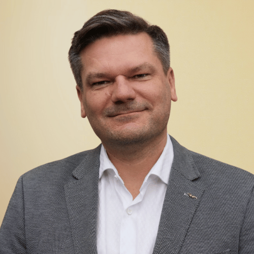 Porträtfoto eines Mannes im grauen Sakko und weißem Hemd vor hellem Hintergrund, freundlich lächelnd.